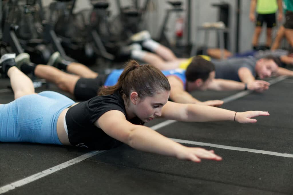 Groupe de personnes effectuant des supermans sur un tapis de gym. La femme au premier plan, concentrée, lève bras et jambes.