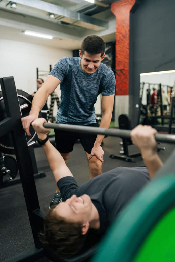 Un spotter souriant aide un homme à faire du développé couché en salle de sport. Ambiance conviviale et soutien mutuel à la musculation.