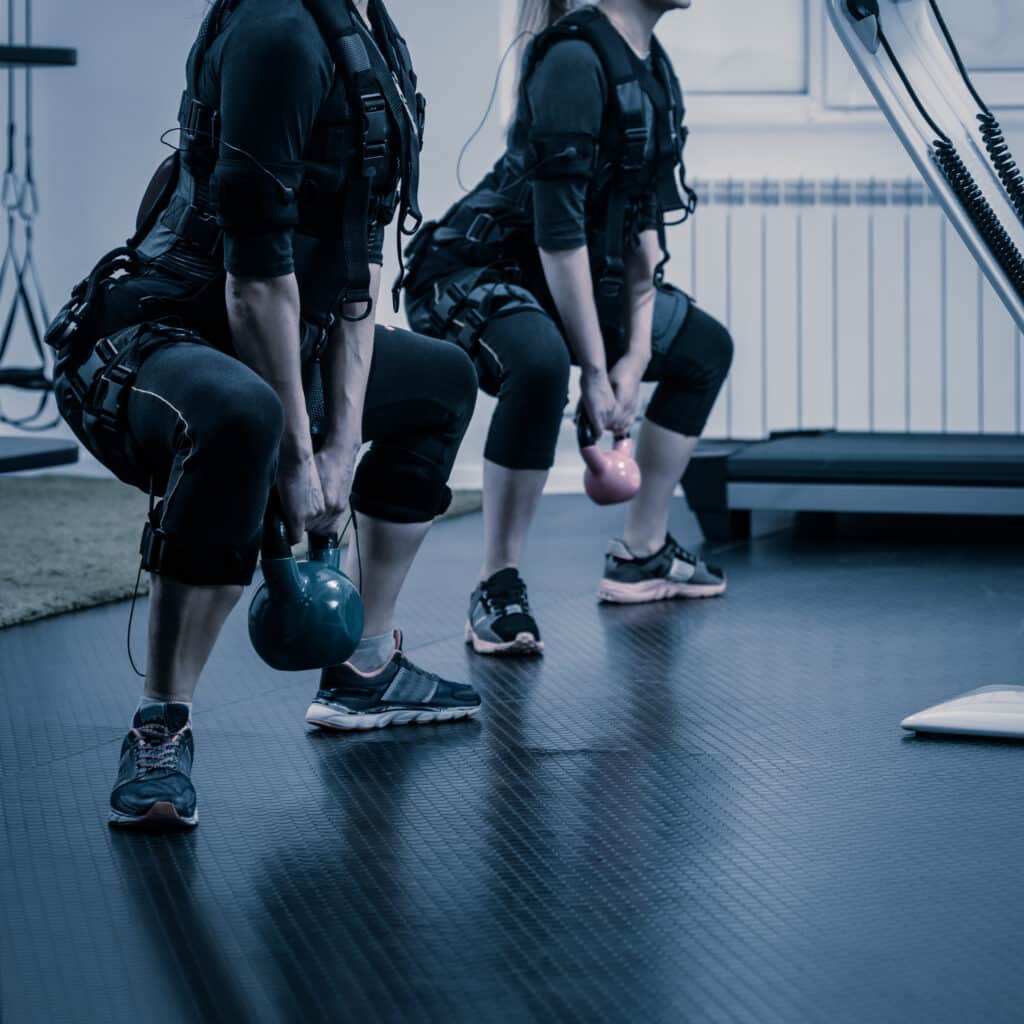 Deux femmes en tenue d'électrostimulation (EMS) effectuent des squats avec kettlebells vertes et roses dans une salle de sport.