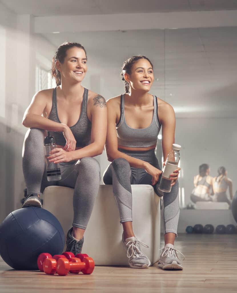 Deux amies souriantes en tenue de sport se reposent au gym après l'entraînement, s'hydratant avec des bouteilles d'eau près d'équipements. Texte: 'MORE WA'.