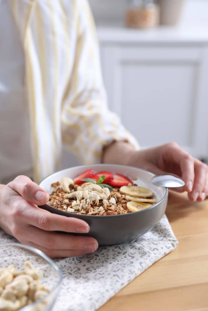 Person tient un bol de granola sain avec yaourt, noix de cajou, fraises, bananes séchées et menthe. Cuillère sur une serviette à motifs.