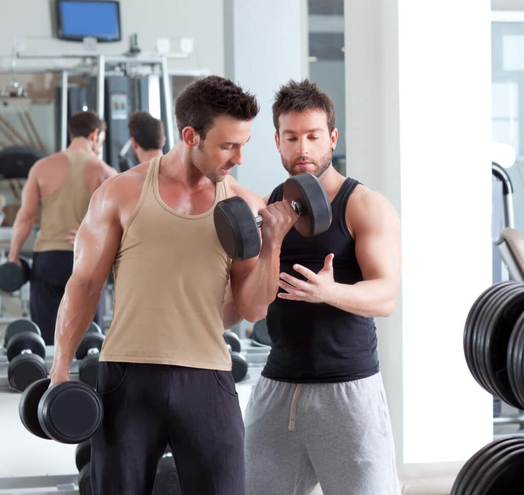 Homme musclé en sueur faisant des curls avec haltère, coach sportif attentif l'encadrant en salle de sport.