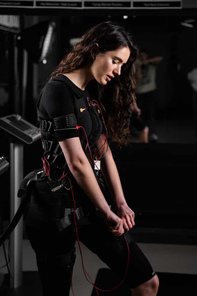 Femme aux cheveux longs en tenue EMS noire à fils rouges, ajustant son équipement. Regard concentré vers le bas. Logo 'mi' sur la cuisse. Salle de sport.